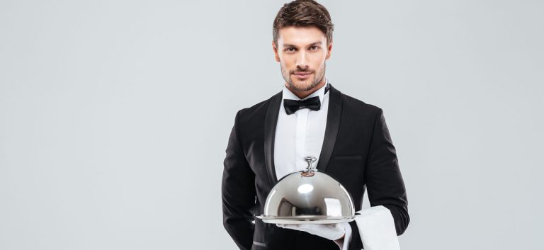 Attractive young waiter in tuxedo holding serving tray with metal cloche and napkin
